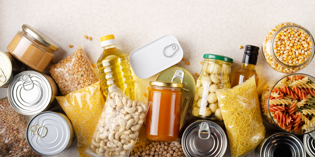 A variety of nonperishable food items, including canned goods, pasta, grains, nuts, cooking oil, and jars of preserves, arranged neatly on a light countertop.