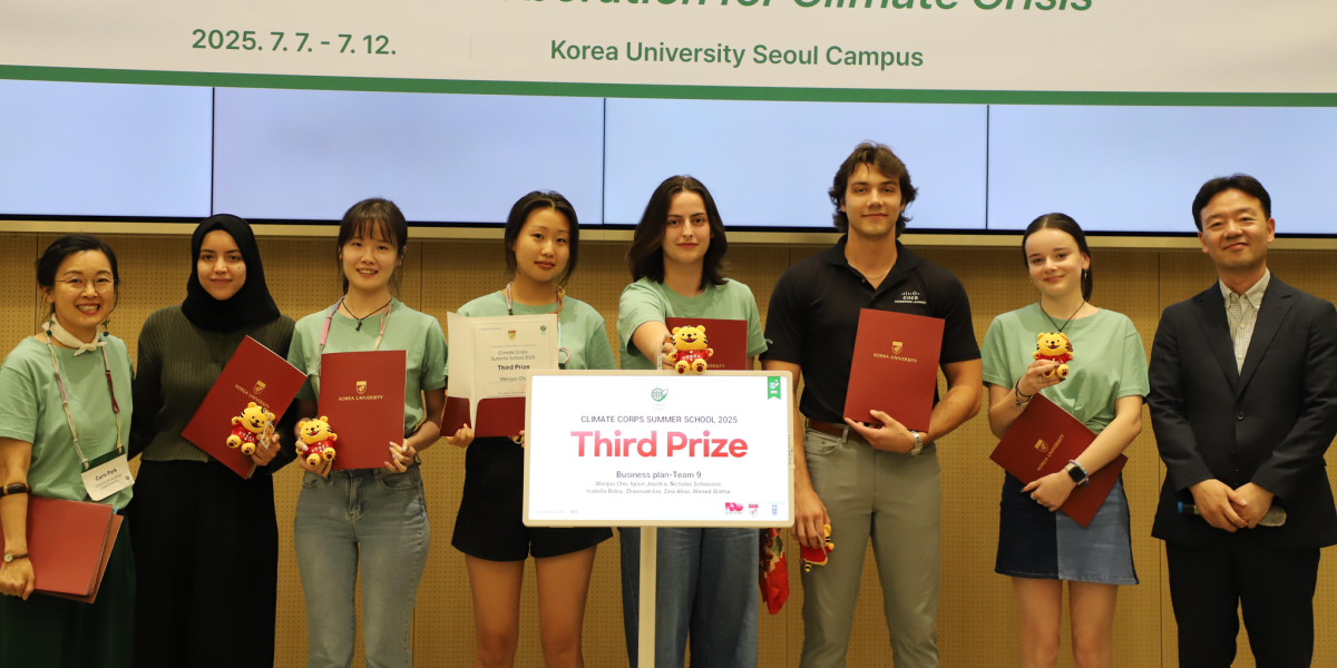 A group of students and faculty stand together on a stage at Korea University Seoul Campus during the 2025 Climate Corps Summer School. Several students wear matching green shirts and hold red Korea University folders and small teddy bear mascots. At center, they display a sign reading “Third Prize – Climate Corps Summer School 2025” for a business plan team. Information science student Nicholas Schiavone stands among the group holding a red folder.