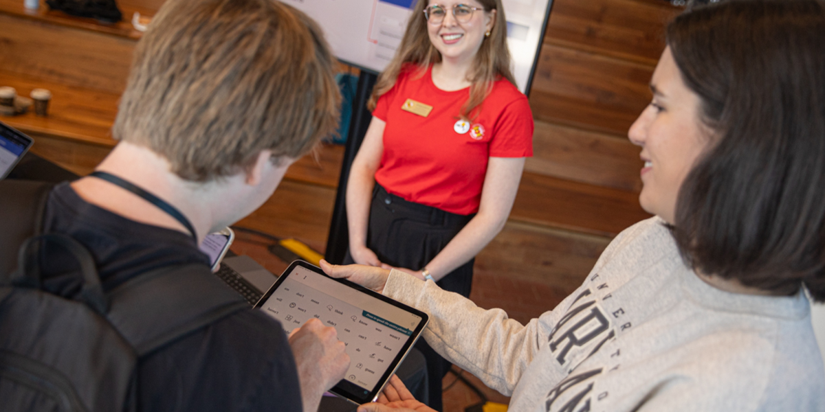 A person demonstrates an AAC (Augmentative and Alternative Communication) app on a tablet while two others observe, one smiling and wearing a red shirt with a name tag. The interaction appears to take place at a research or technology event.