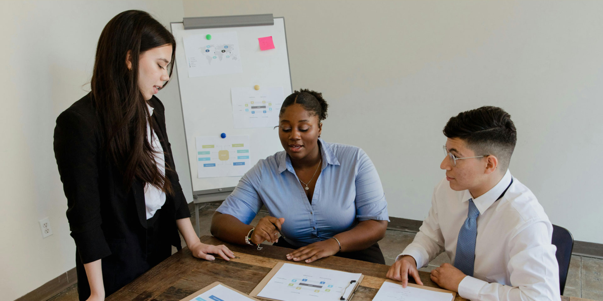 Three students in professional attire collaborate at a table with printed charts, while a whiteboard with diagrams stands in the background.