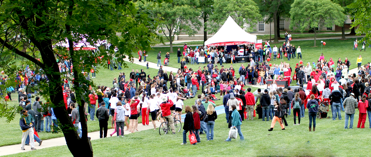 A wide-angle, high-angle view of a large, diverse crowd of students and visitors gathered on a lush green lawn at the University of Maryland campus. In the background, white event tents and classic red-brick collegiate buildings are visible under a clear sky.